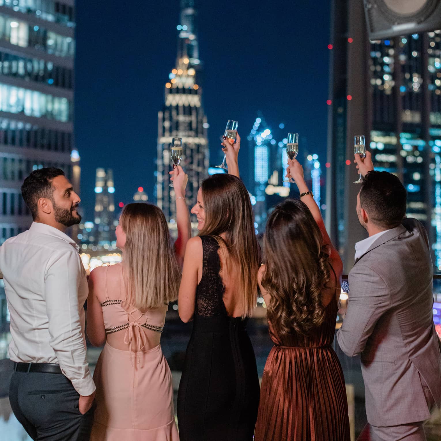 Three women wearing party dresses, one man wearing a grey suit and another man wearing a white dress shirt and dark pants stand with their back to the camera, raising their glasses to the nightime Dubai skyline