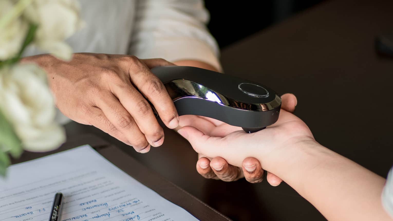 A person scans another's palm with a smooth, shiny, black electronic device beside a completed medical form on the table..
