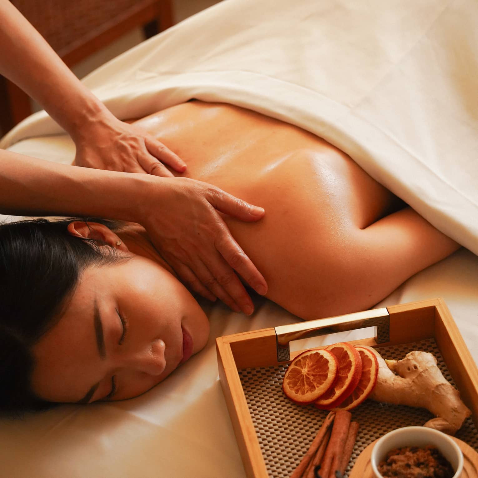 A woman gets a massage on a spa table, near a tray of dried orange and cinnamon sticks