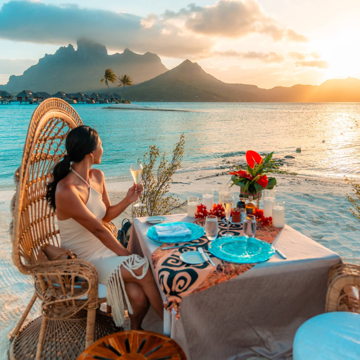 Person in high-backed wicker chair sits at table on beach, holding glass of white wine and gazing at the sunset