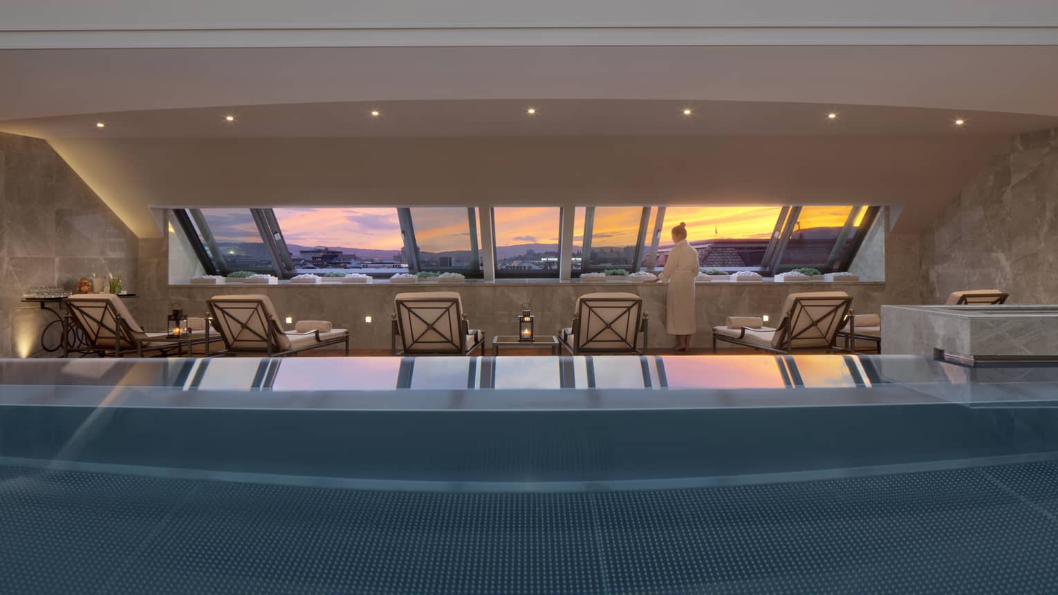 Woman in robe stands on deck beside blue indoor swimming pool, lounge chairs, looking at sunset through window