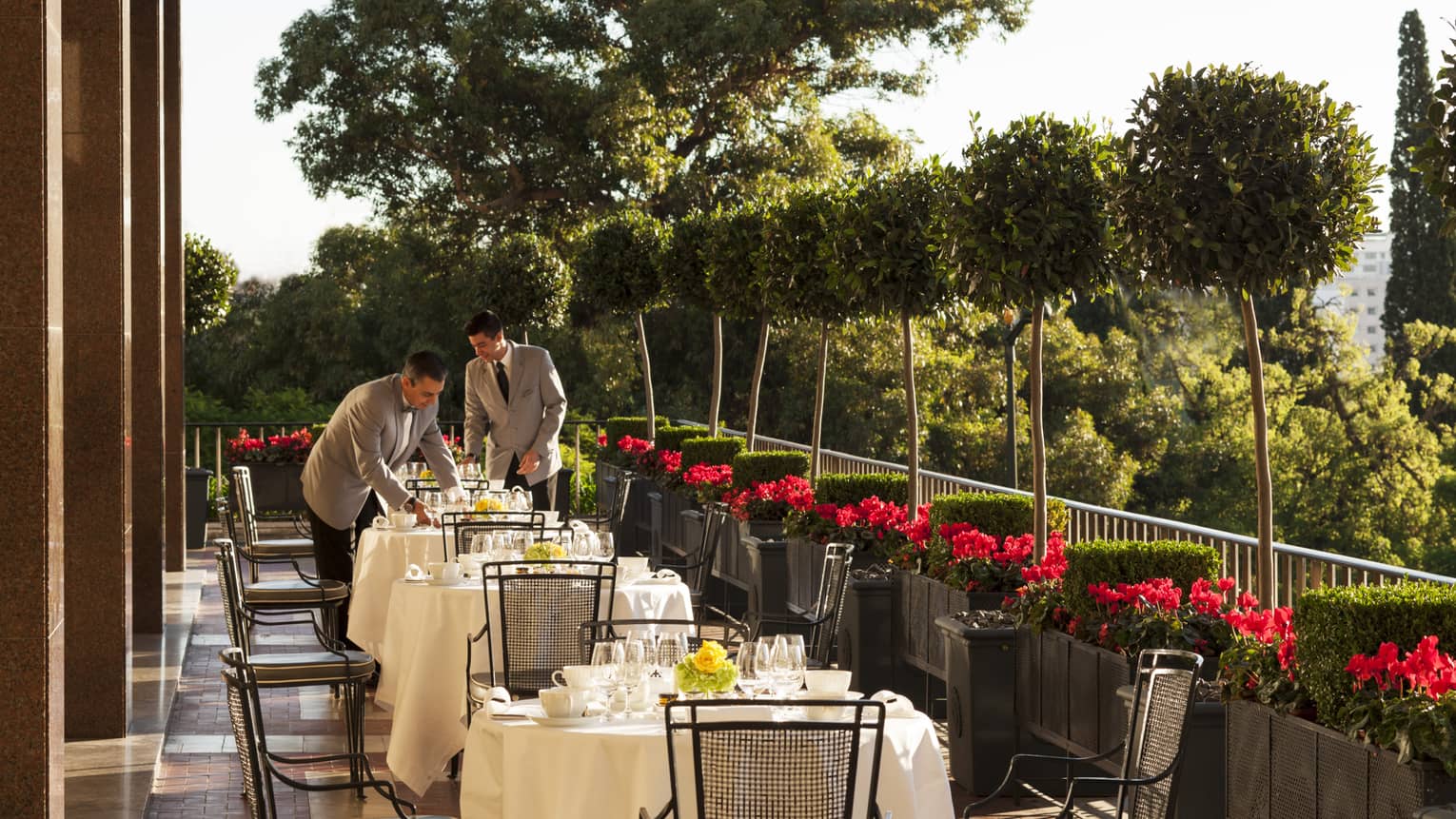Two servers in grey jackets with black ties set outdoor dining tables with white tablecloths, dishes, red flowers along side