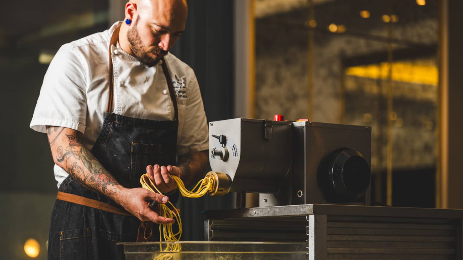 A chef catches fresh long pasta noodles as they emerge from an industrial pasta-making machine in a warmly lit kitchen.