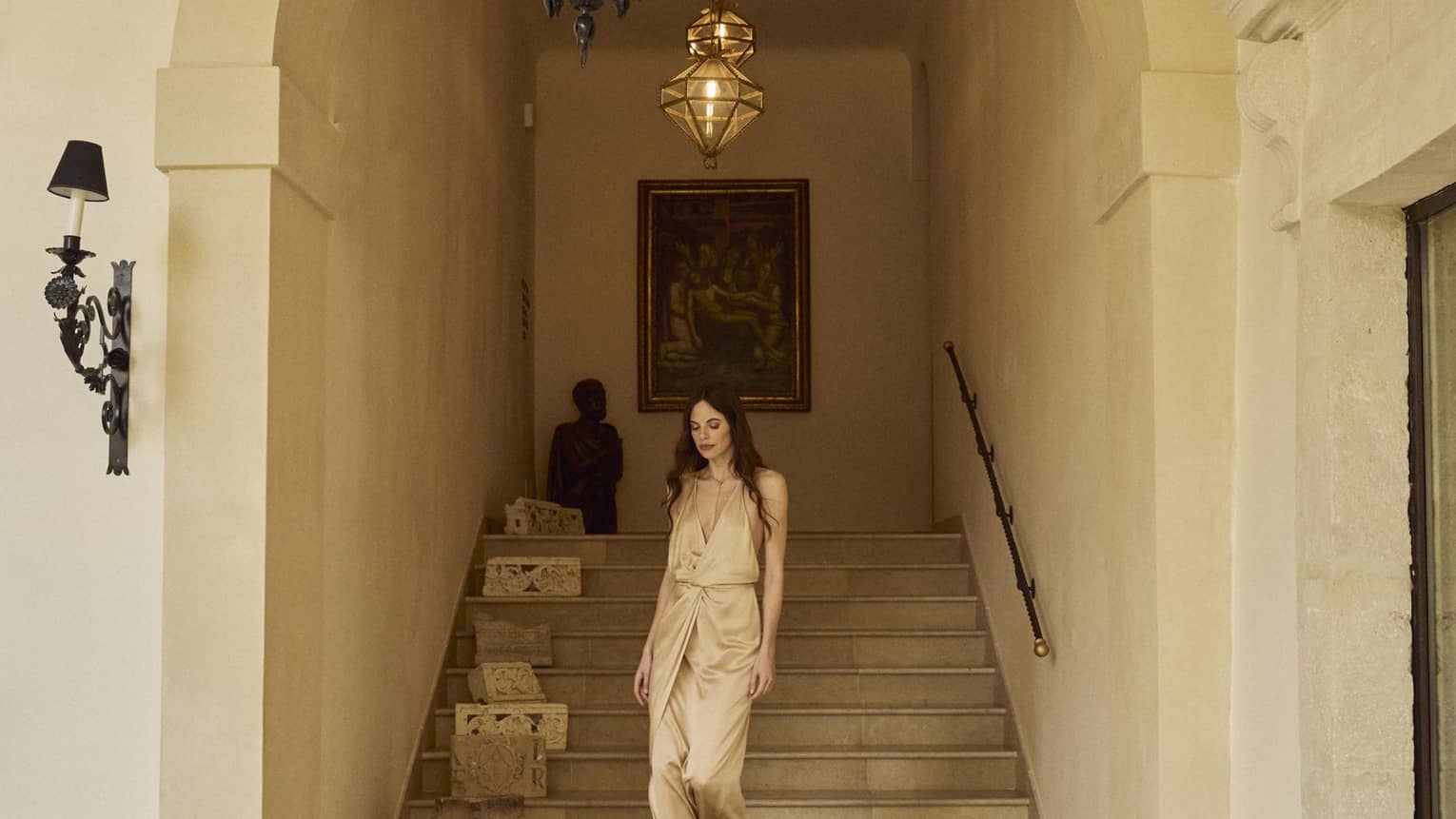 Woman wearing Champagne-coloured satin dress walks down the stairs in an arched walkway