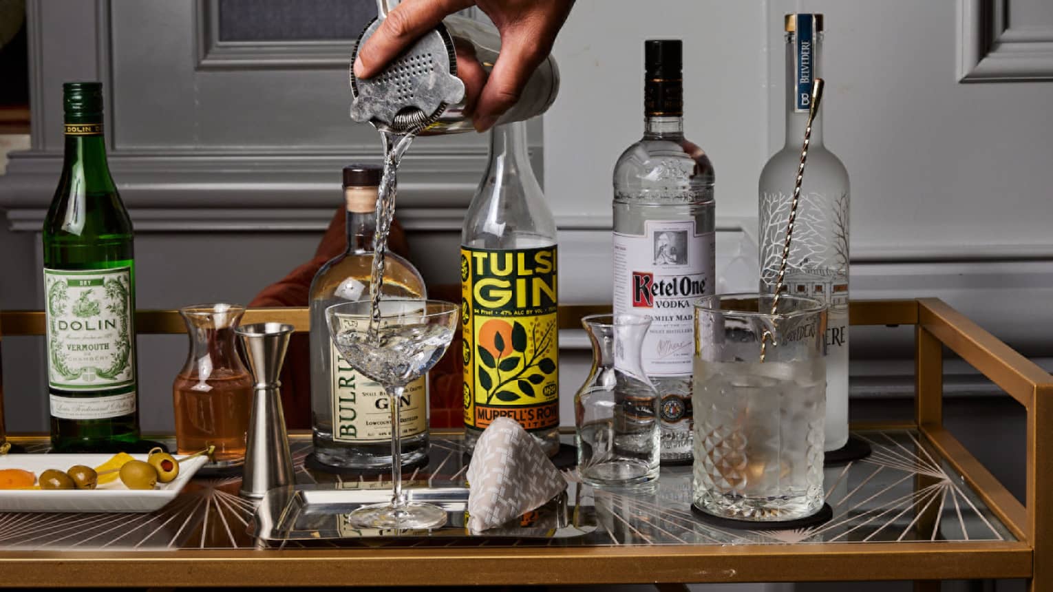 A bartender pours from a shaker into a coupe glass next to bottles of gin, vodka and vermouth, a jigger, carafes and olives.