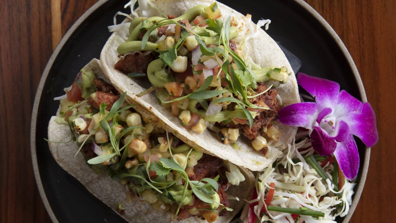 Aerial shot of two brisket tacos with pineapple salsa and orchid garnish on a black ceramic plate