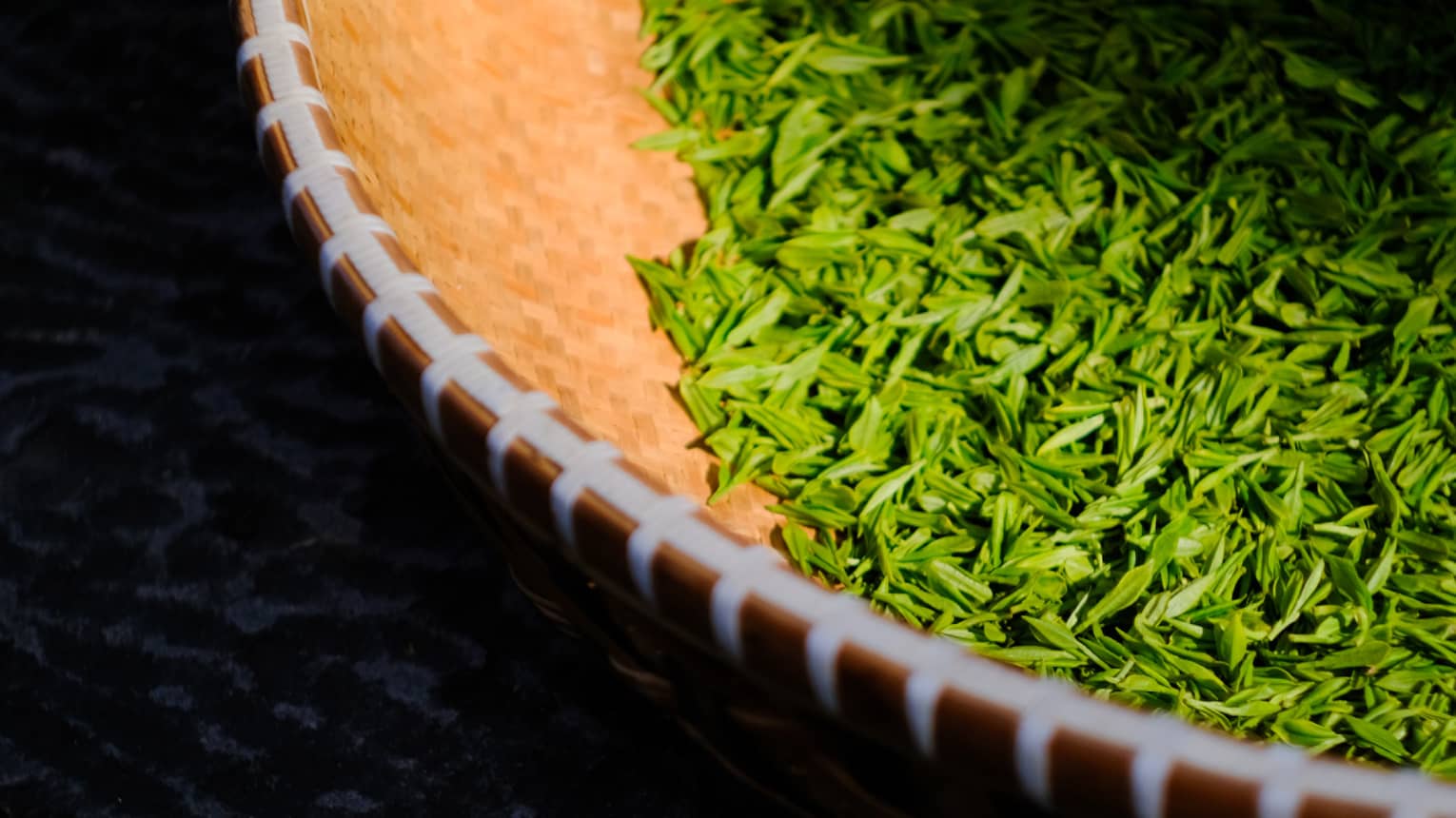 A tightly-woven straw-coloured basket with a white rim holds a small pile of vivid green tea leaves.