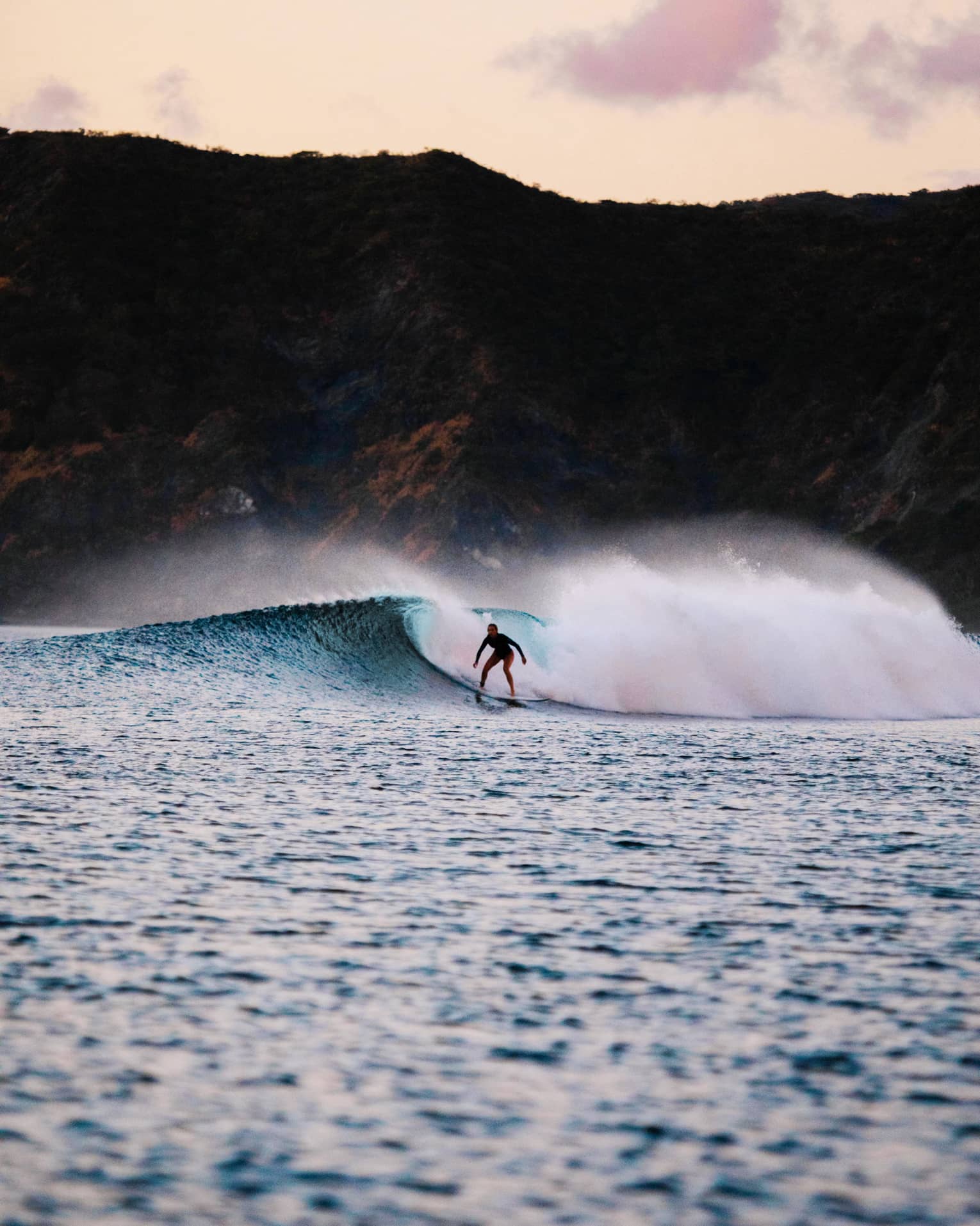 A surfer rides a wave amid a great cloud of frothy sea foam; a jagged cliff in the background, pink and yellow skies above.  