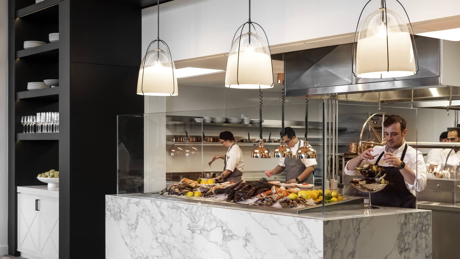 At an elegant marble counter, a server arranges an impressive seafood presentation. Behind, chefs at work in the kitchen.