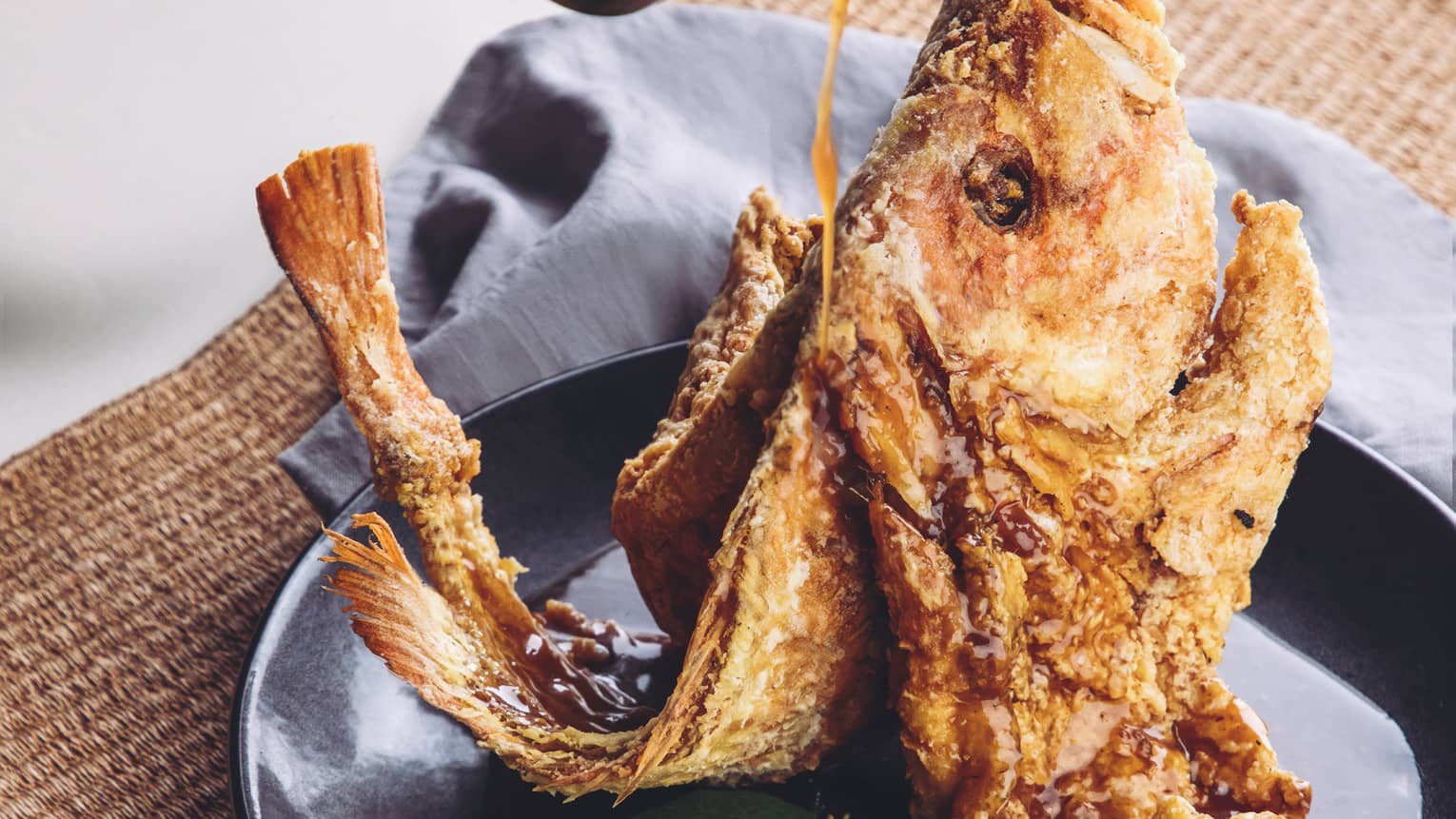 Staff member pours sauce on a whole fried fish artfully displayed on a black ceramic dish with fresh herb garnishes