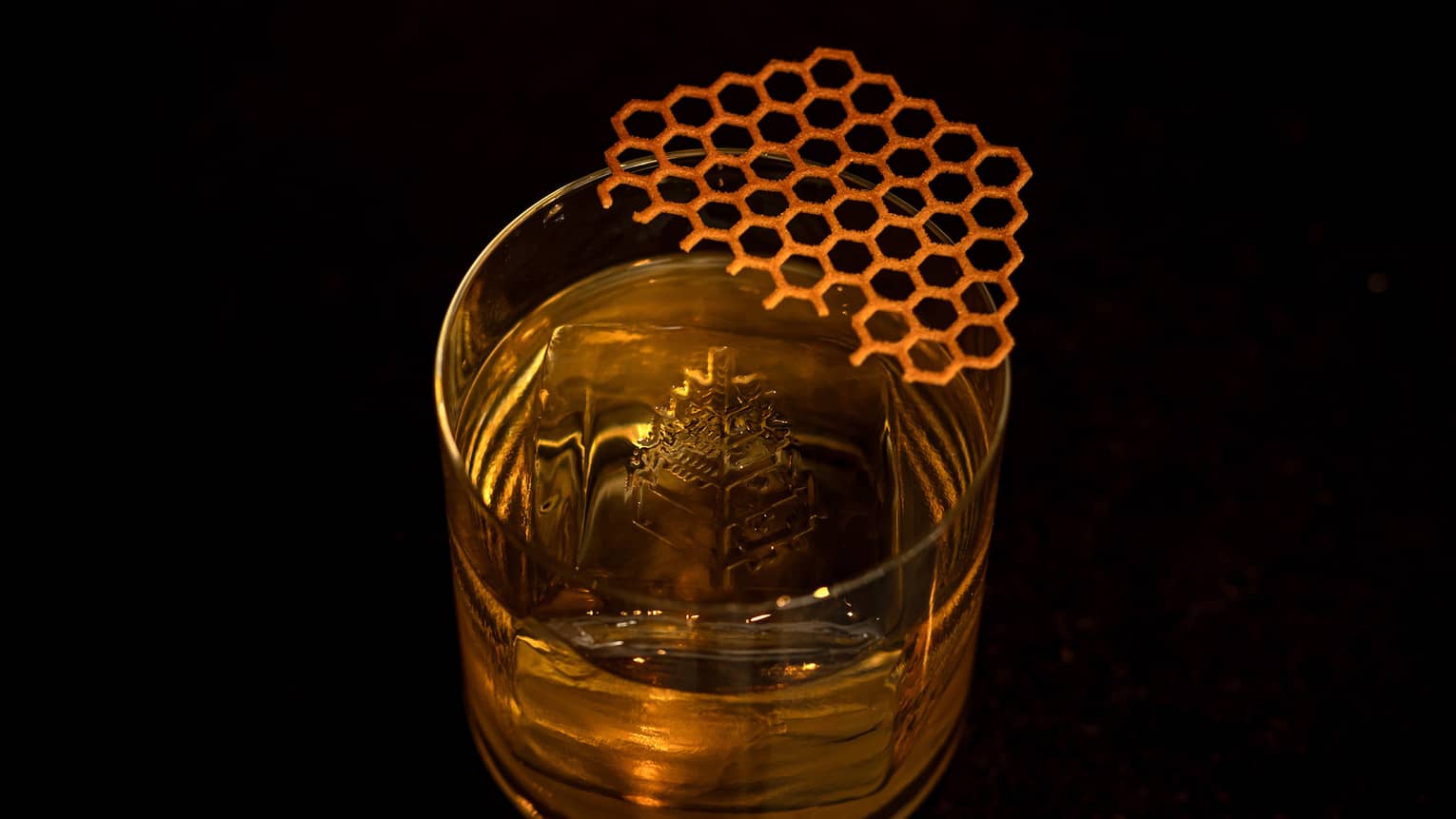 A clear tumbler contains a golden-hued cocktail, ice and garnish