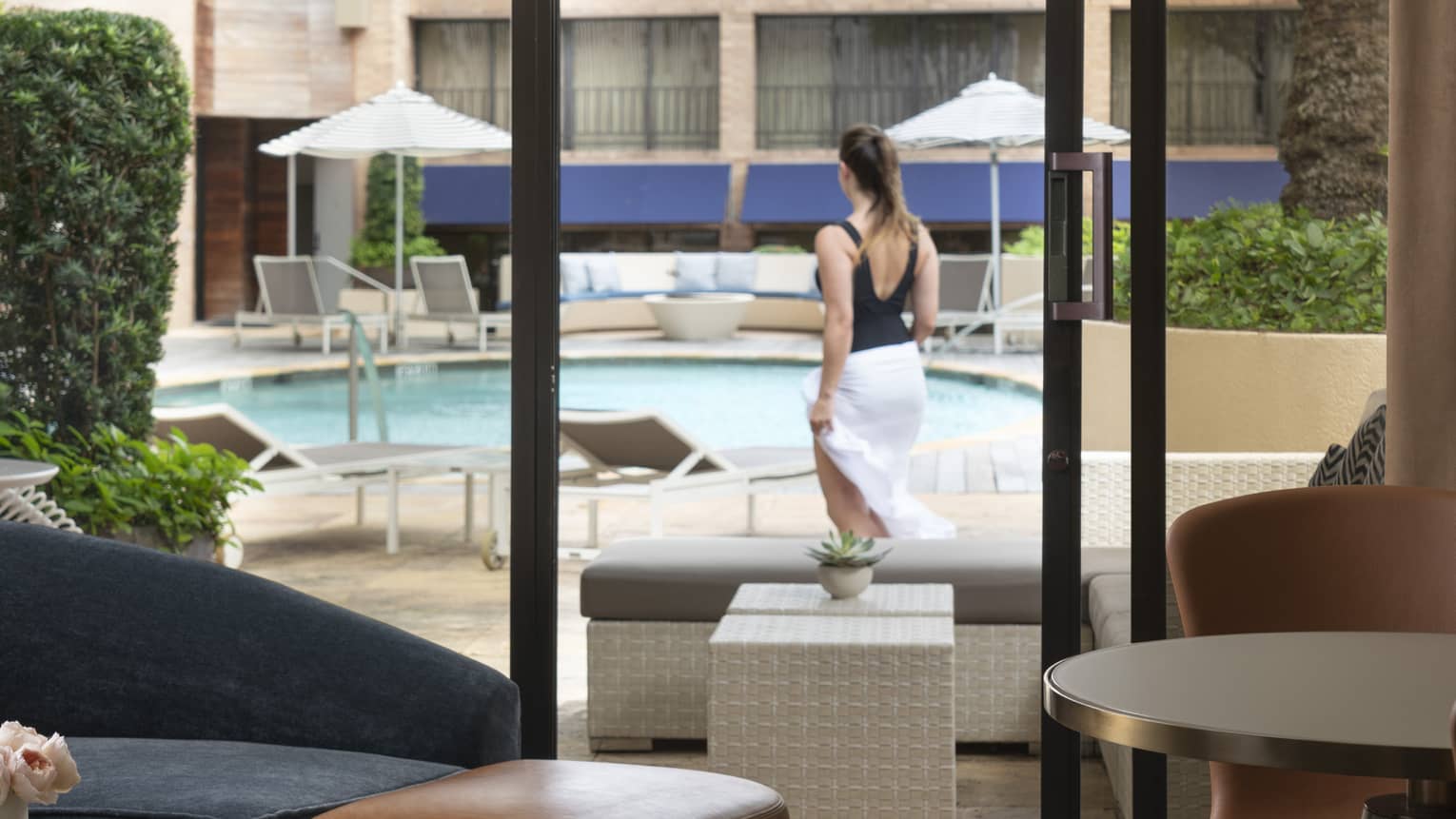 An indoor cabana decorated with modern furniture; outside the glass door a guest surveys the outdoor pool and lounge chairs.