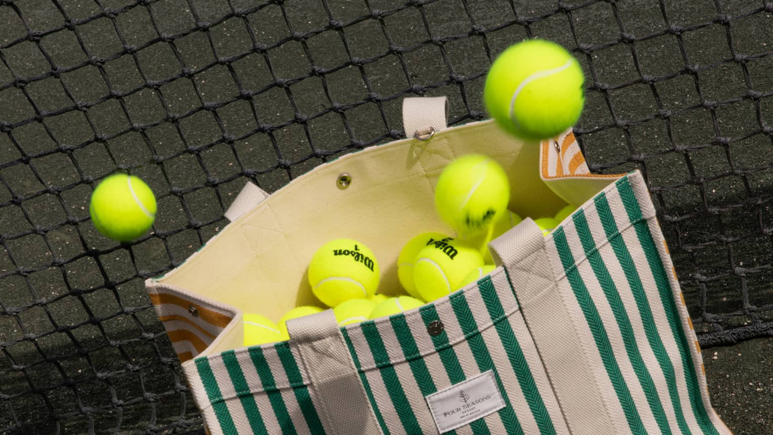 Green-and-white striped canvas bag filled with tennis balls leans against a net