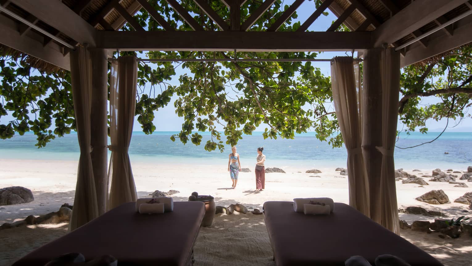 Couples massage beds under cabana in front of two women walking on white sand beach