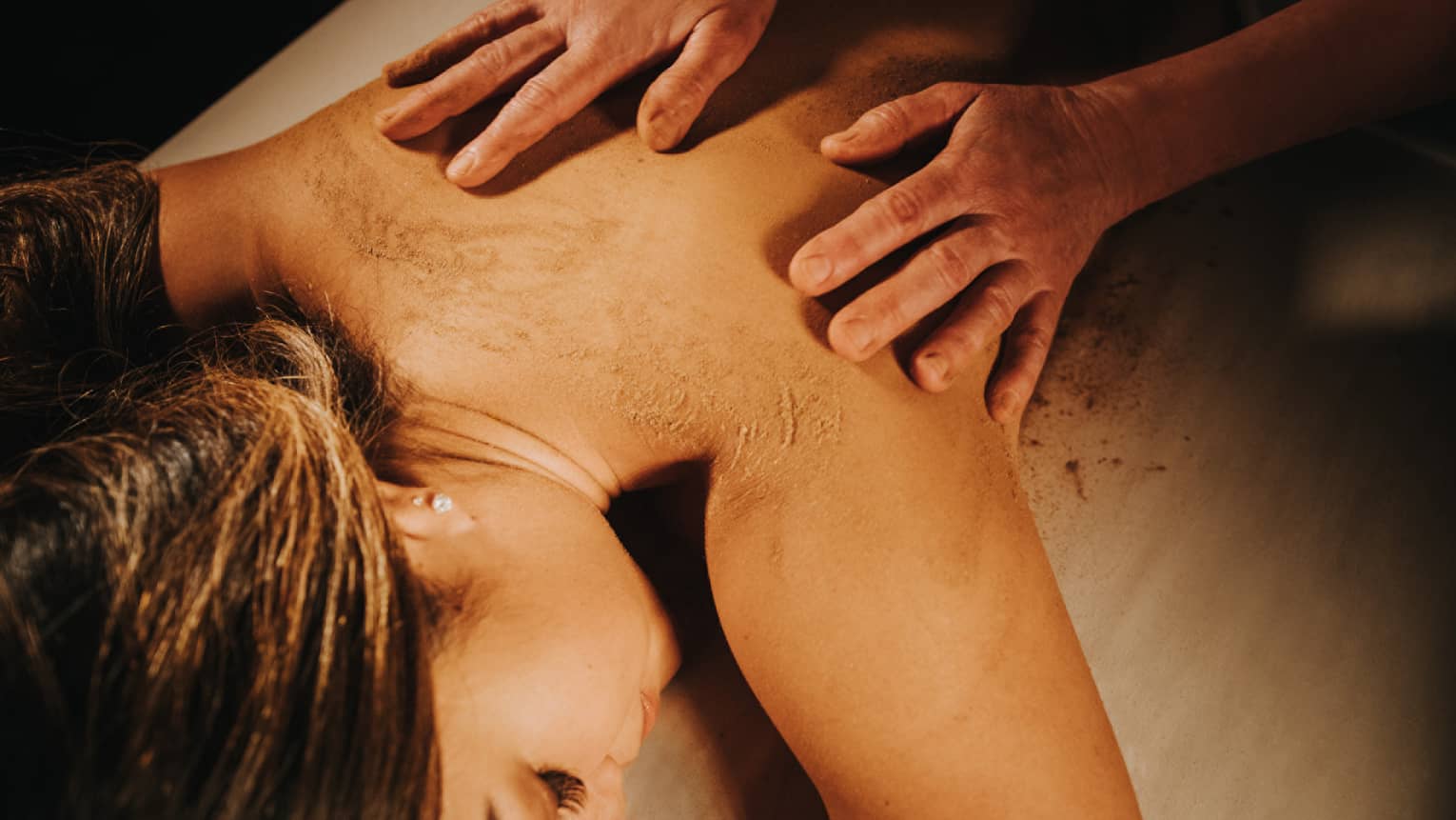 Two hands on woman's bare back as she lays on massage table in dimly lit room