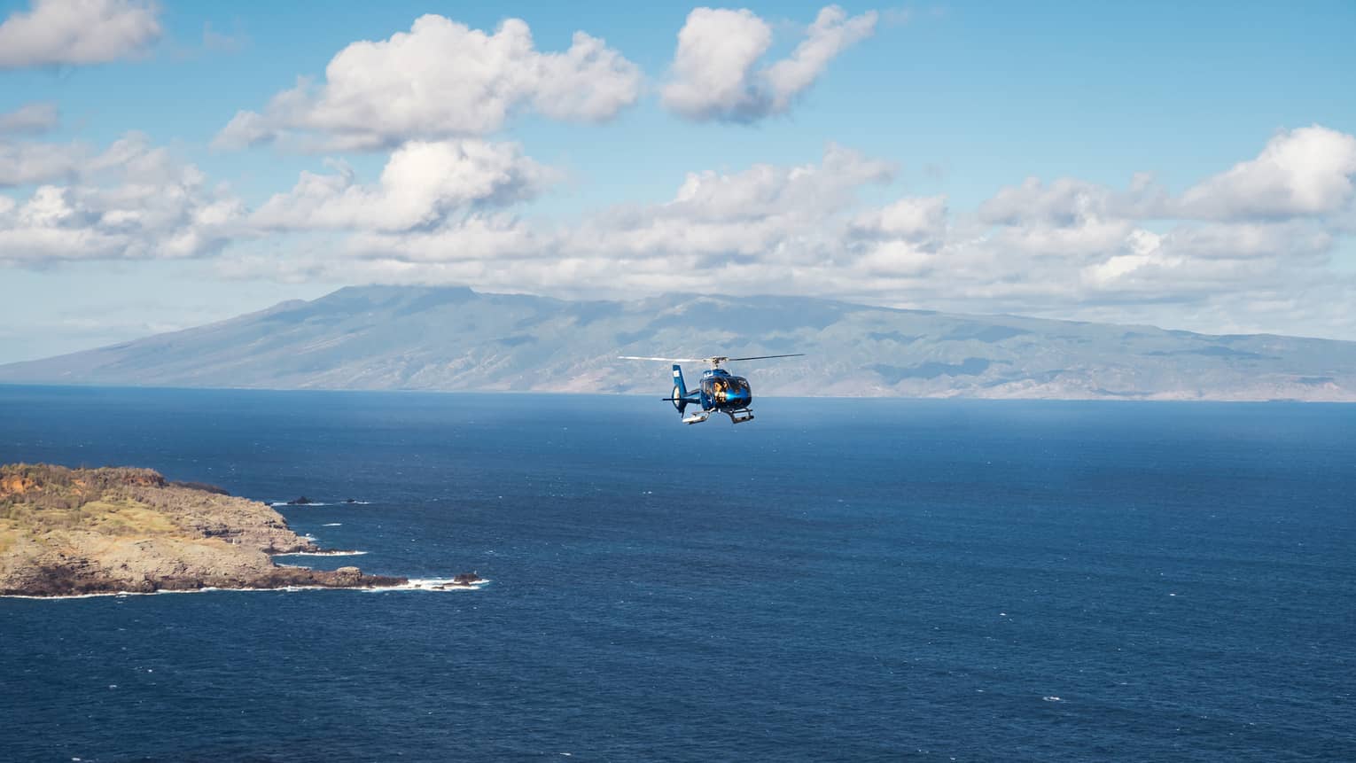 A helicopter flies over open ocean with a mountainous island and blue sky dotted with clouds in the background