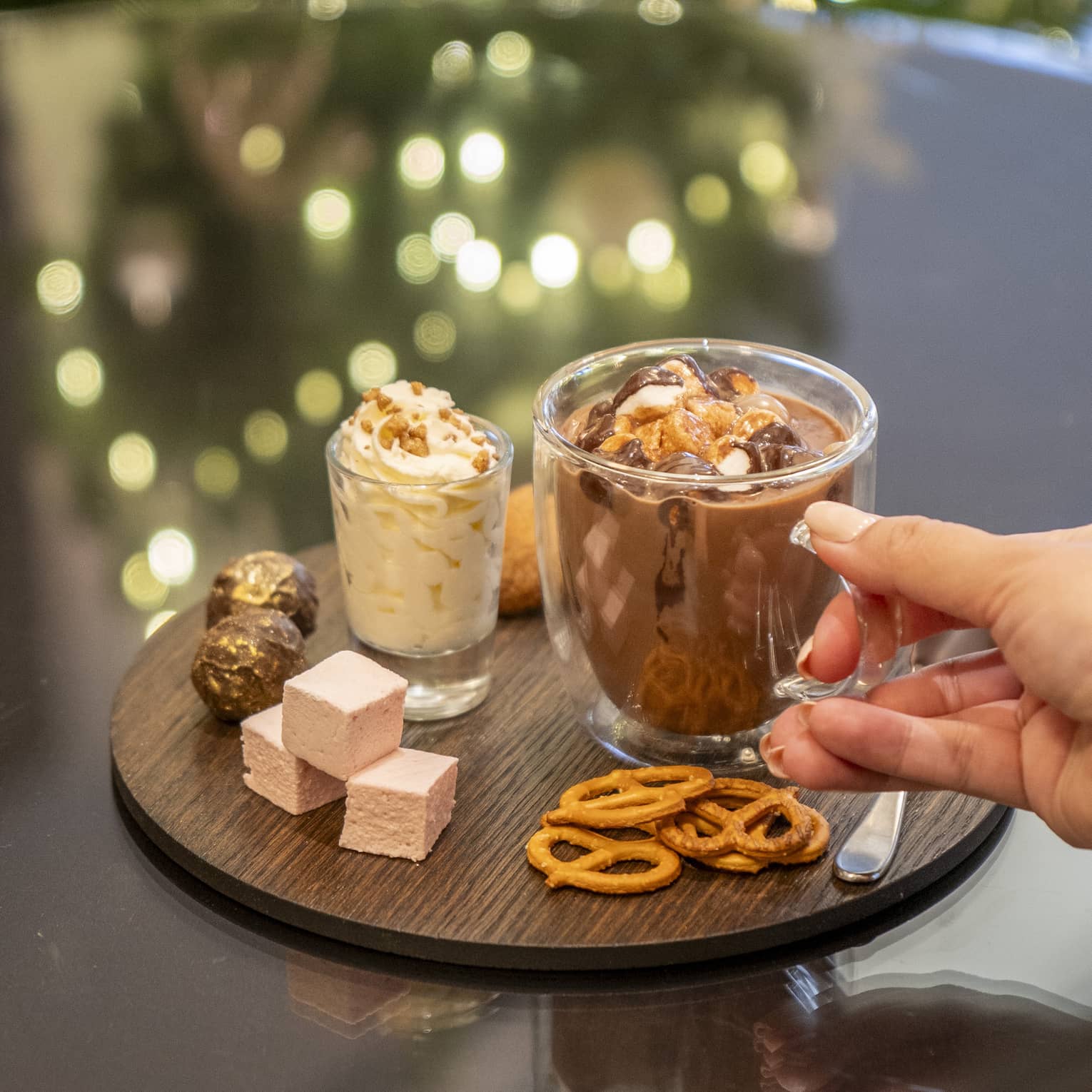 Round wooden platter with a glass mug of hot chocolate, small glass of whipped cream, thre pink marshmallow squares, pretzels and assorted chocolates