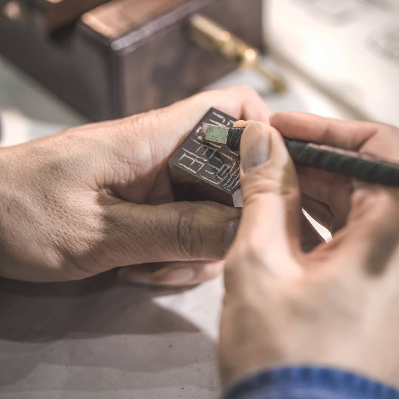 A hand holds a small block of dark stone while another hand carefully uses a tiny chisel to carve symbols into its surface.