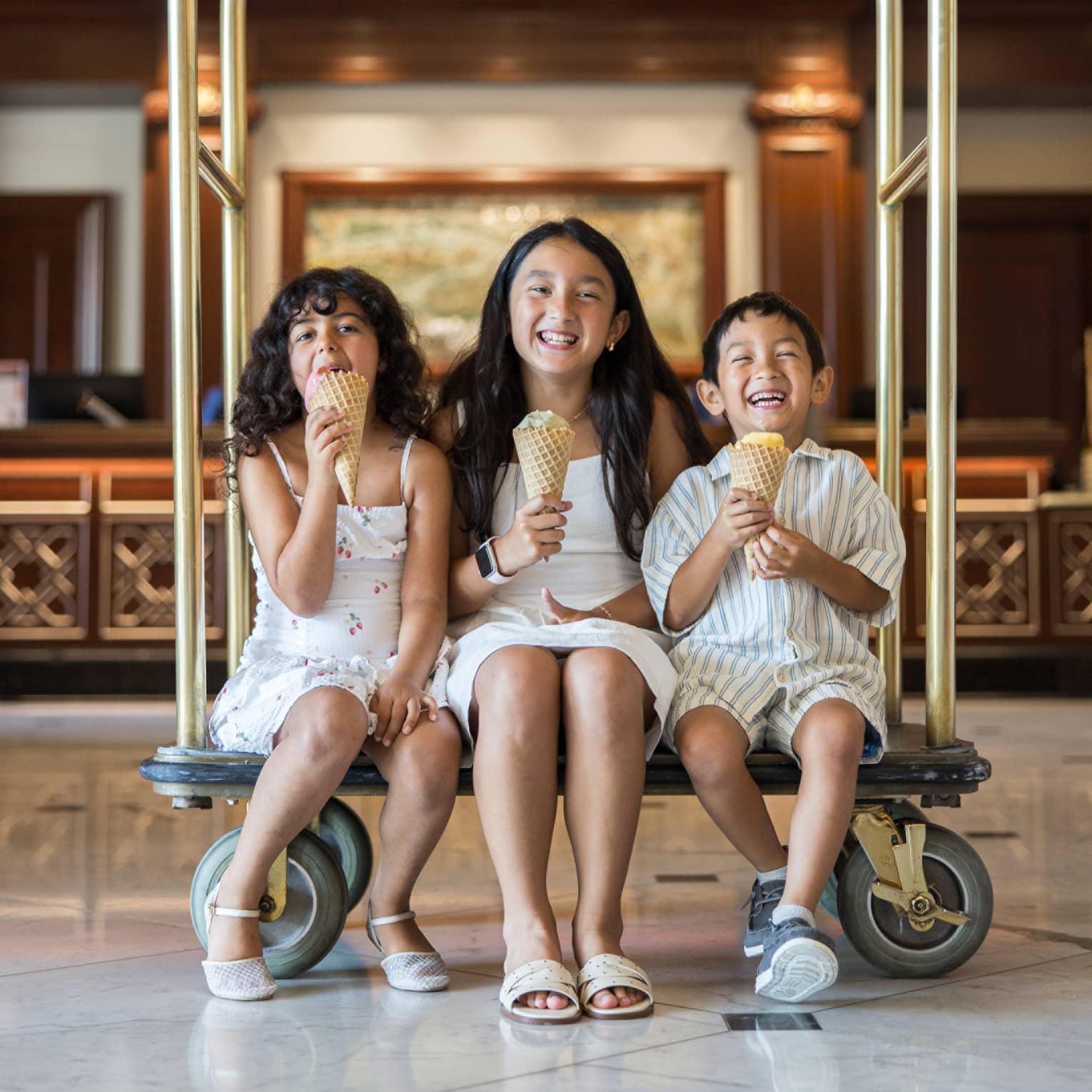 Three children sit on a gold hotel luggage cart eating ice cream