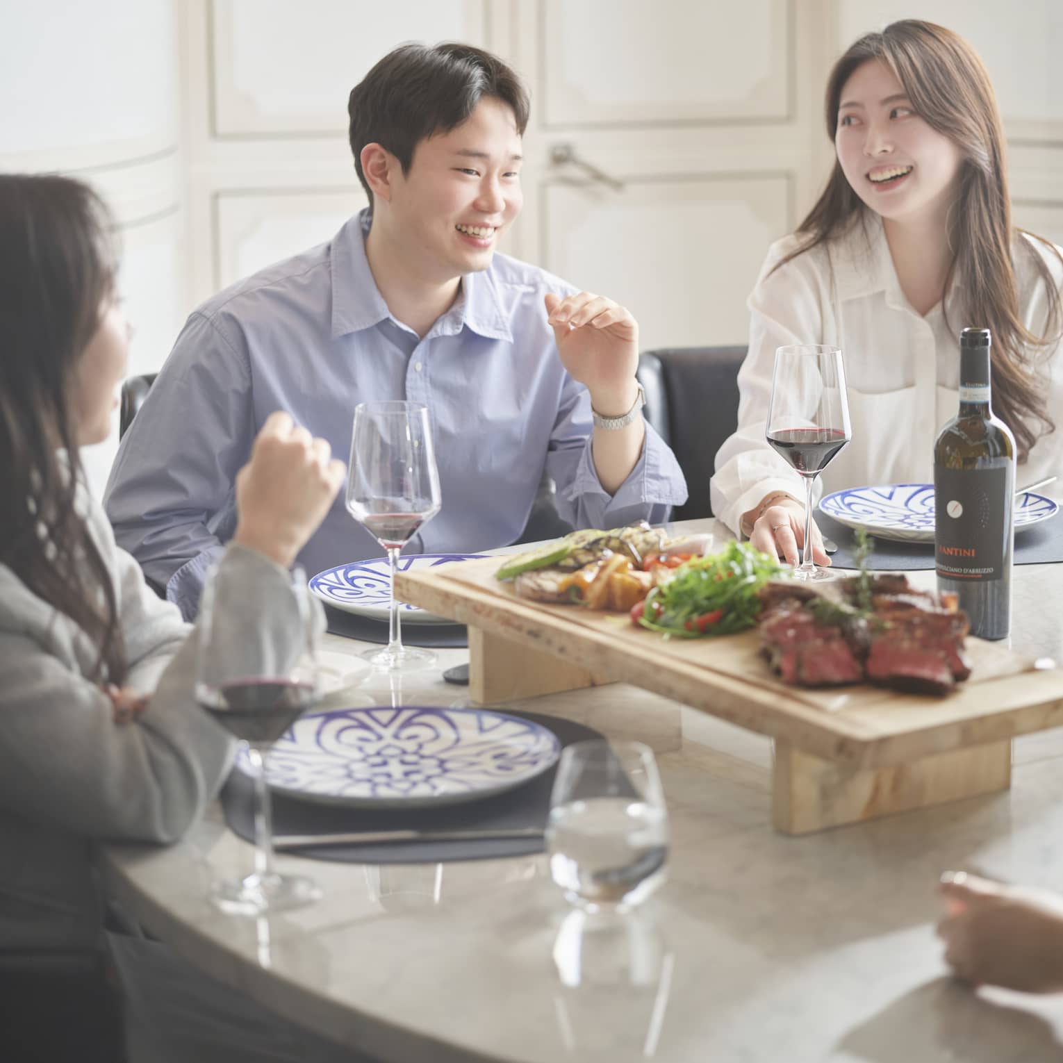 Group of friends sit around a table for a meal together, glasses of red wine and wooden display of meat and vegetables surrounding them