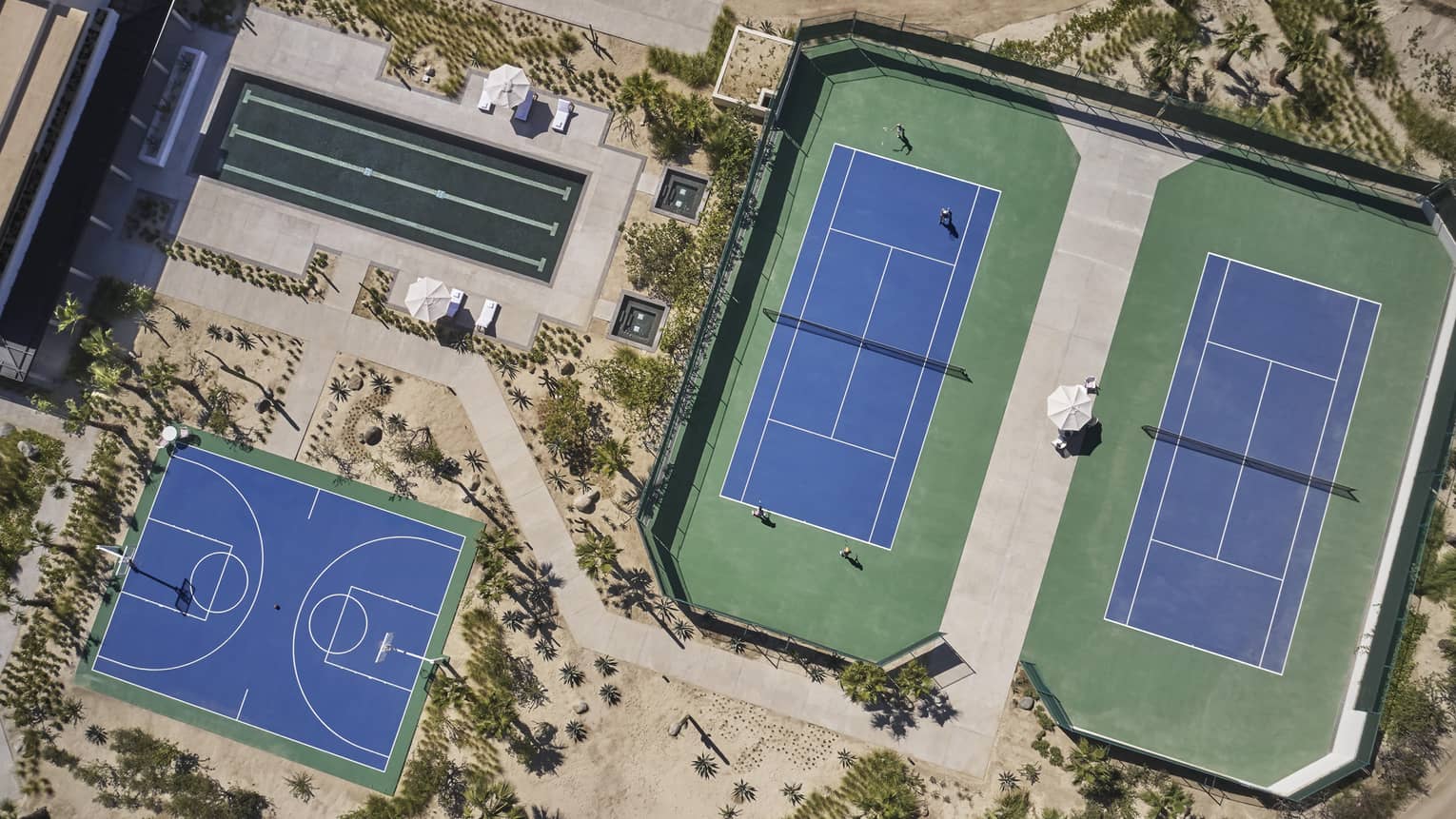 Aerial view of two green and blue tennis courts, one long rectangular pool and one blue basketball court surrounded by sand and greenery