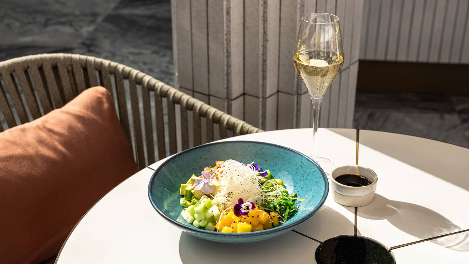 A turquoise bowl on a white table holds a variety of colorful ingredients and is set next to a small container of dark sauce and a pale drink in a clear stemmed glass