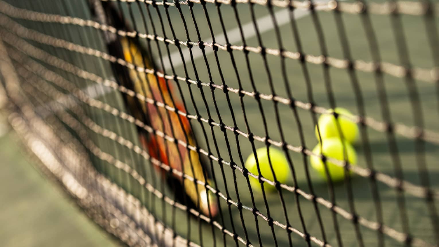 Close-up of tennis racket against net, three green tennis balls on ground