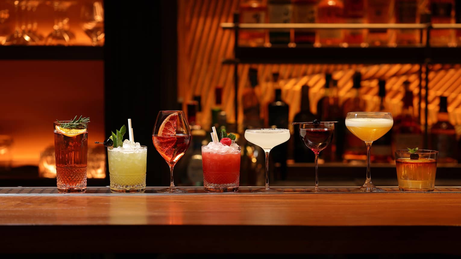 A line of cocktails in various tumblers and stemmed glasses against a backdrop of blurred liquor bottles in reddish lighting.