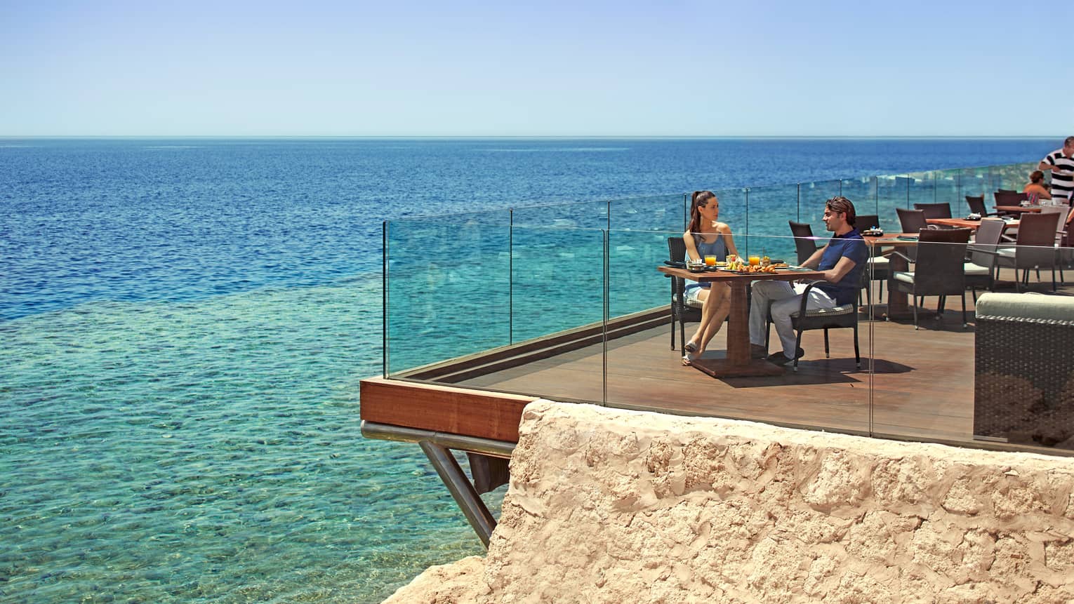 Couple dines at sunny corner patio table by glass balcony overlooking turquoise waters of Red Sea