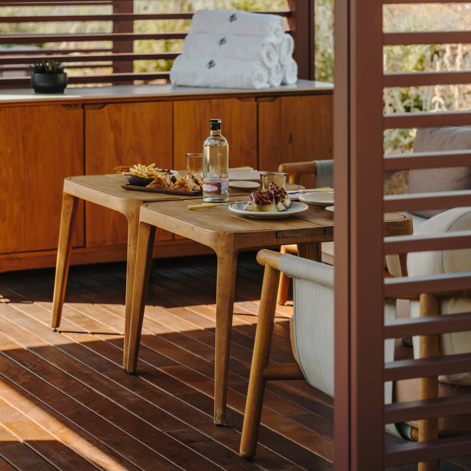 A private pool cabana with small tables, comfortable seating, a cabinet with rolled-up towels and slatted walls admitting filtered light sunlight.