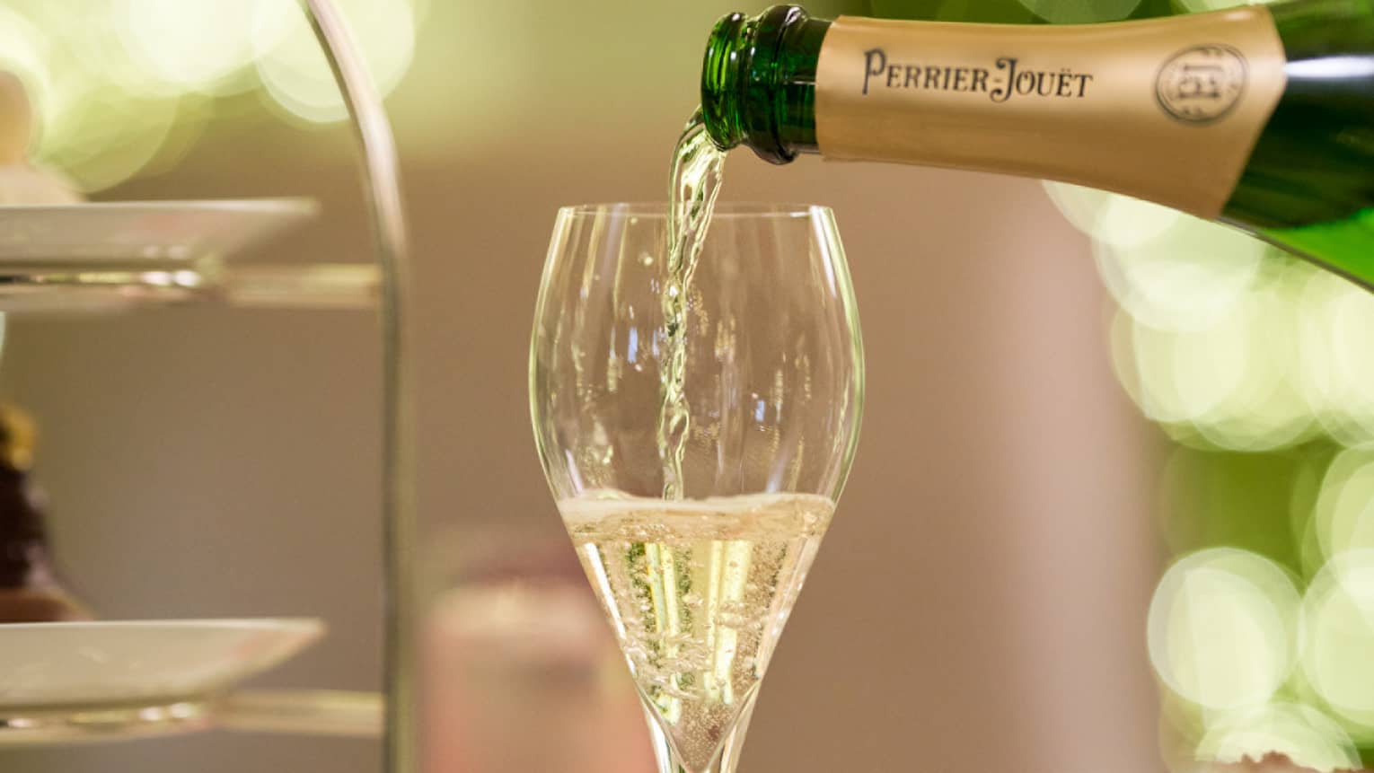 Champagne is poured from a bottle of Perrier Jouet into a glass next to a tiered tray of treats and a frothy cup of espresso.