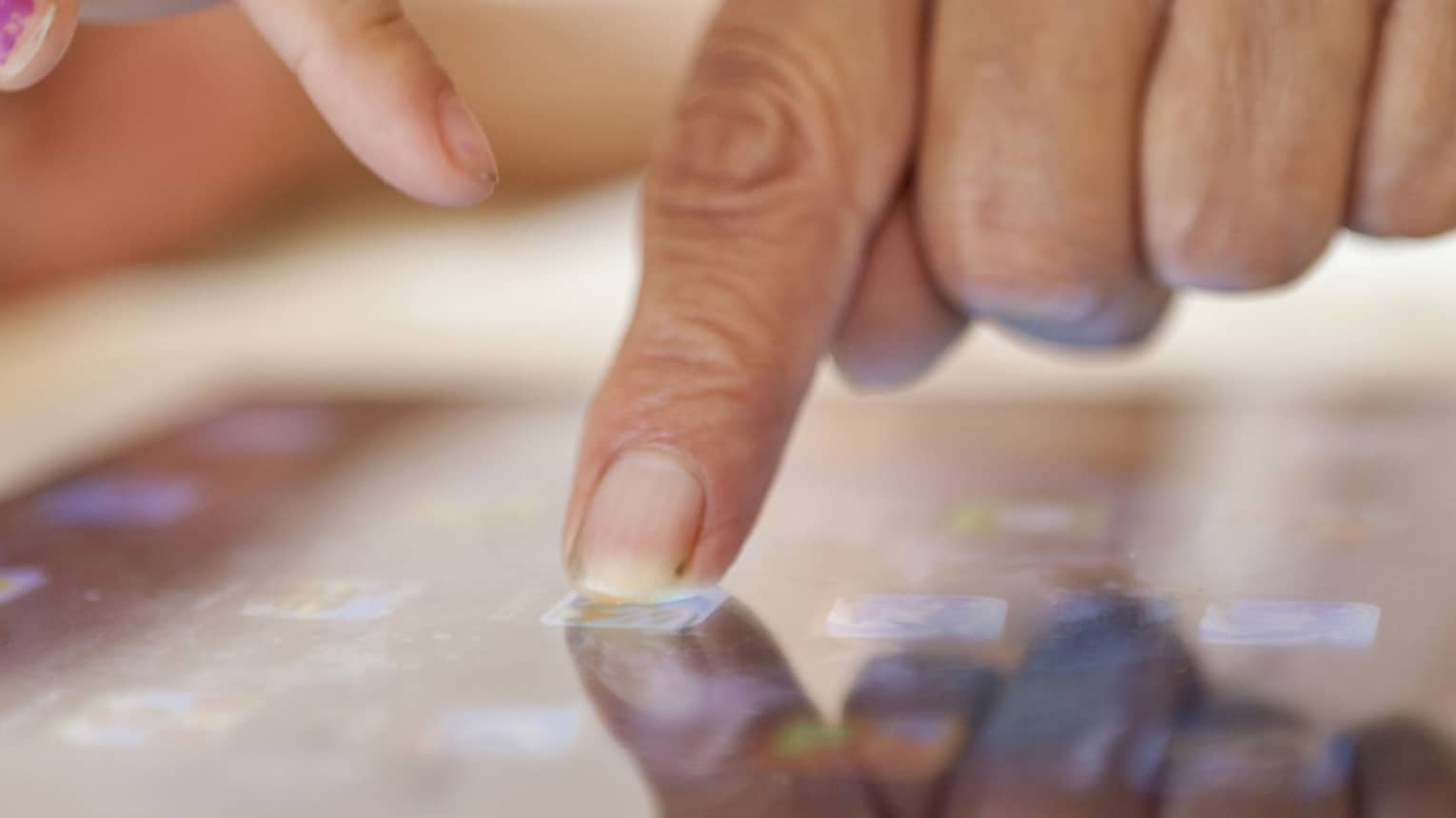 Close up of two hands, one with an index finger pressing a button on a touchscreen and the other pointing at the screen