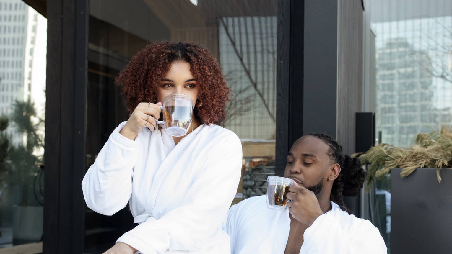 Man and woman drink coffee in white spa robes by outdoor firepit