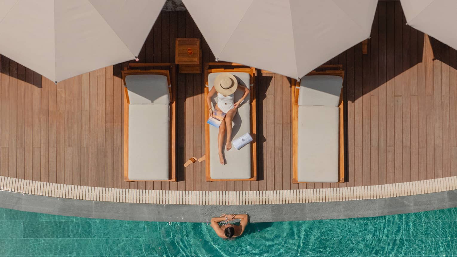 Aerial view of woman wearing white bathing suit relaxing on a lounge chair next to a pool with a man standing in the water with his arms on the ledge