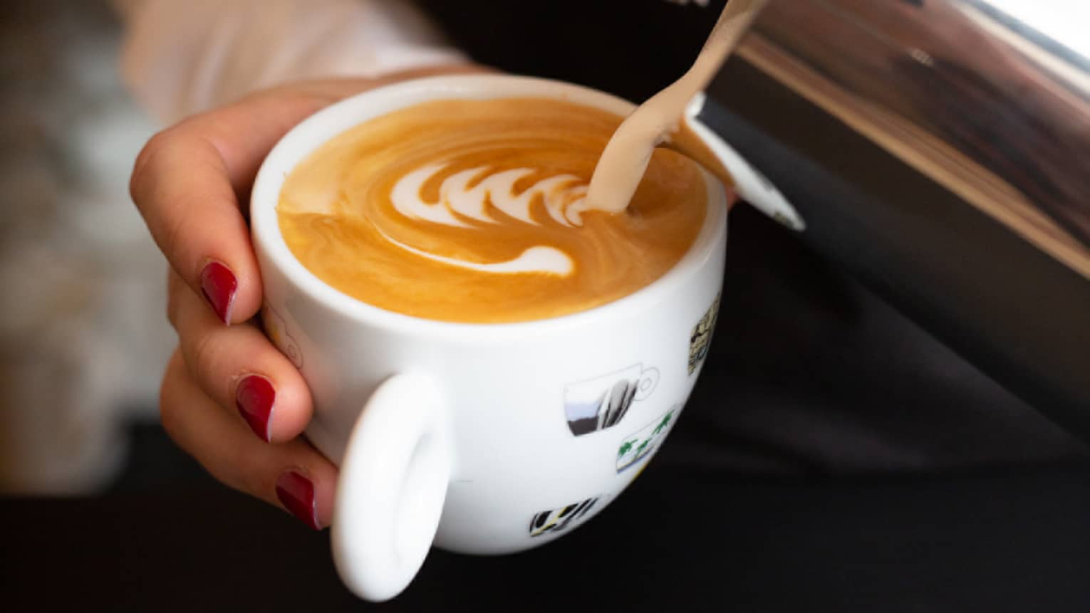 Close-up of hand holding and stirring white mug with foamy latte coffee