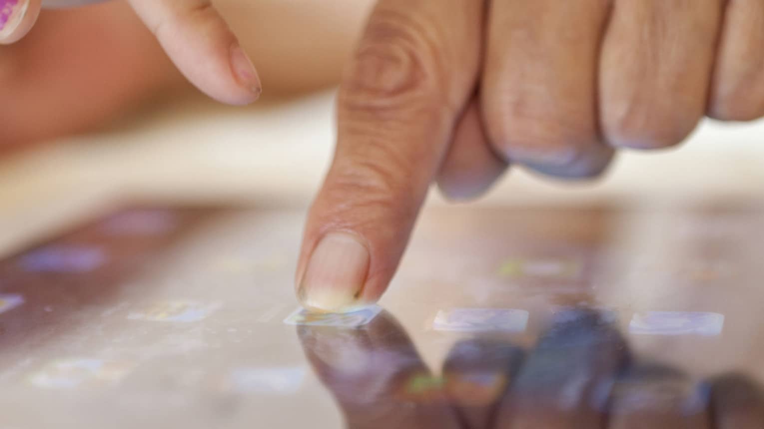 Close up of two hands, one with an index finger pressing a button on a touchscreen and the other pointing at the screen