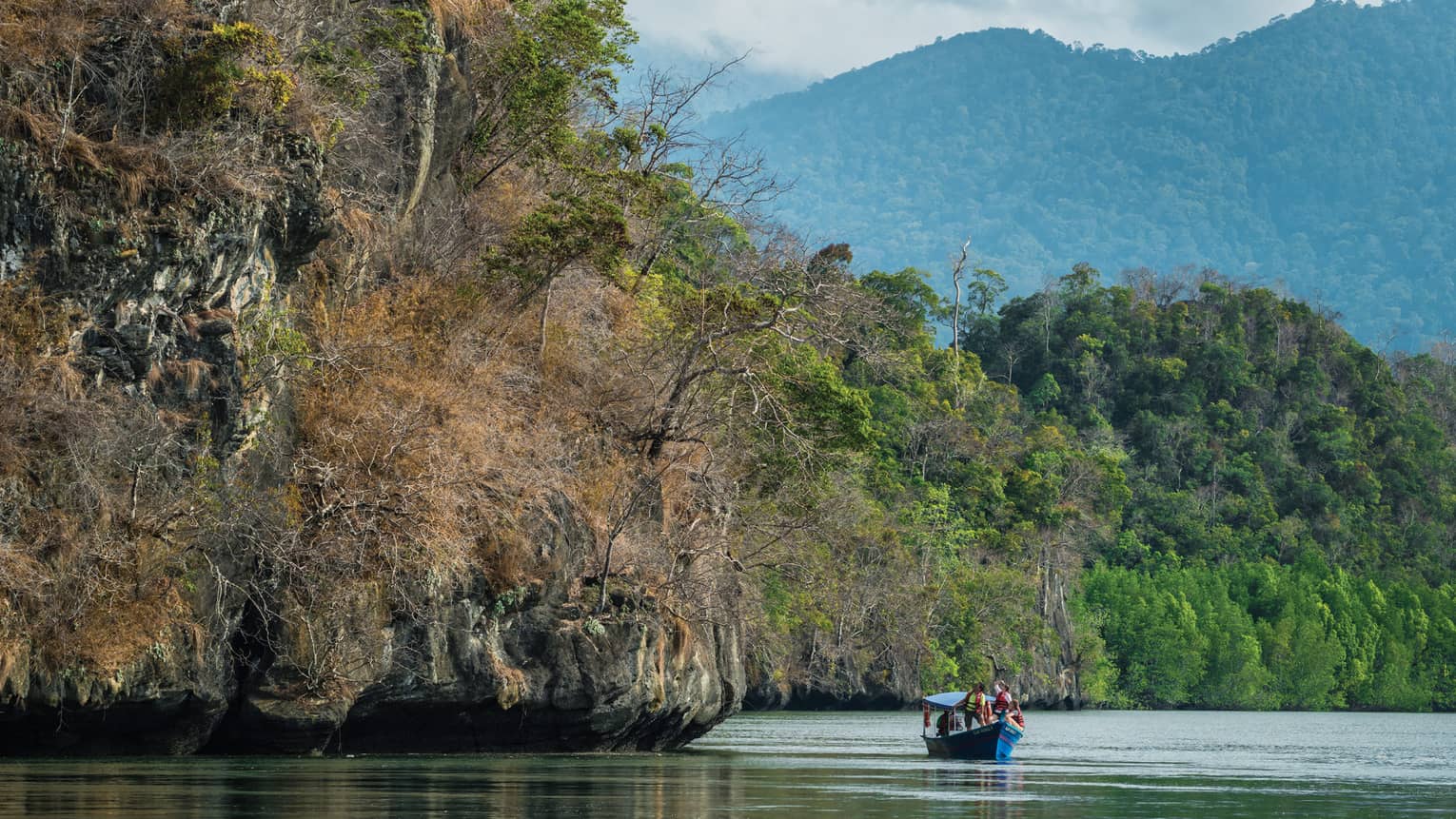 Small blue boat on river under large cliffs, trees