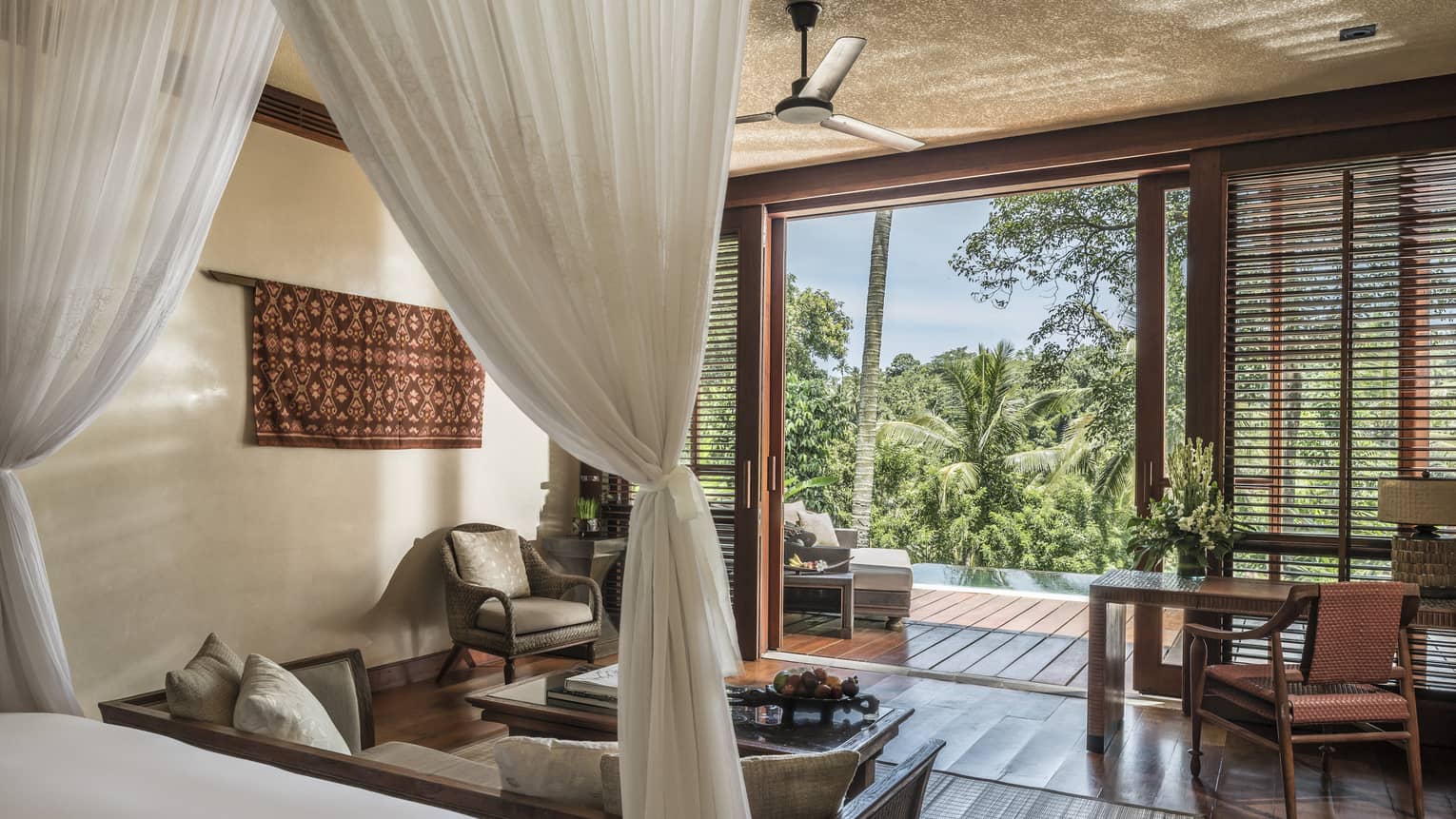Villa bedroom with canopy bed, hardwood floors, desk and desk chair, arm chair, sliding door to pool and balcony