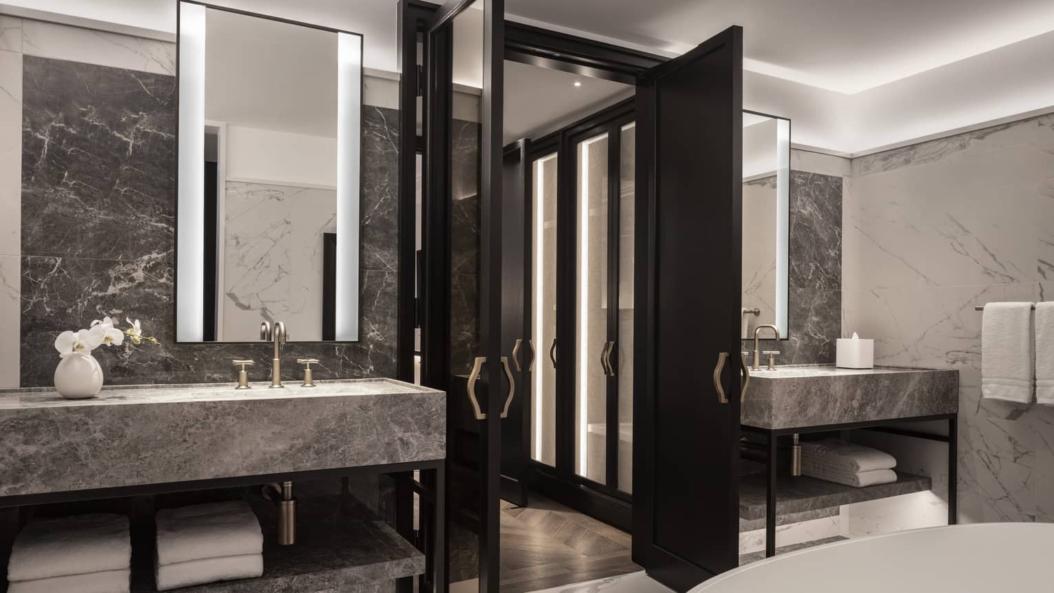 Bathroom with dark marble countertops, backlit mirrors, brass fixtures, double sinks, a freestanding bathtub and folding doors
