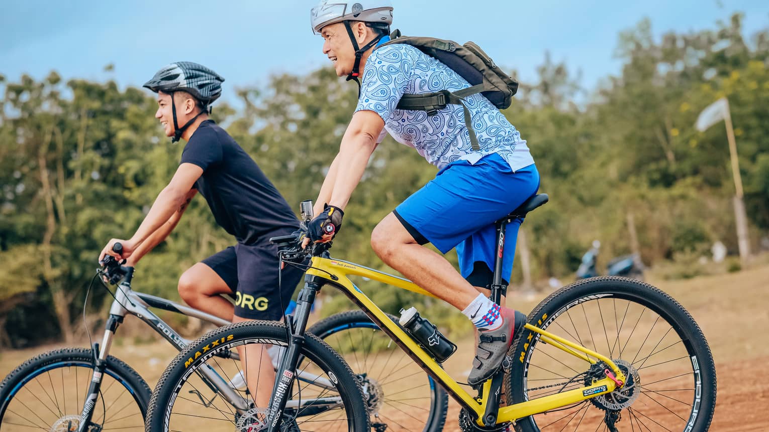 Side view of two smiling mountain bikers in helmets as they coast down a gentle slope on a dirt path alongside trees.