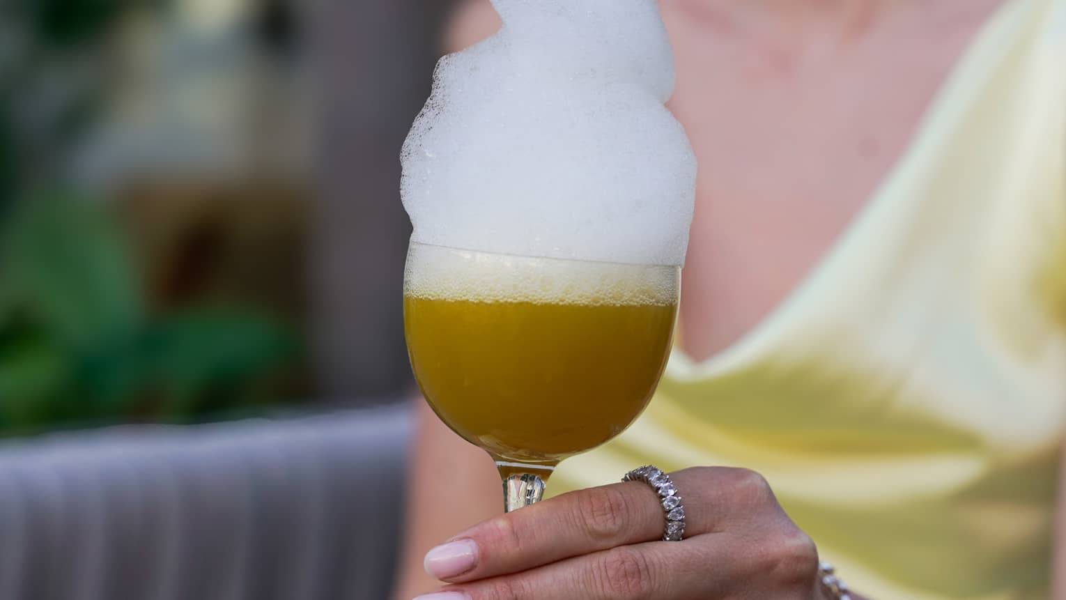 A guest wearing a satin yellow dress holds a stemmed glass filled with a tan-hued beverage topped with an overflowing dollop of foam