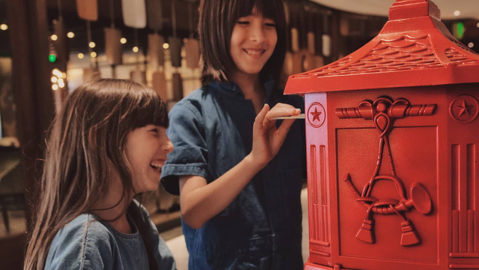 Two children put letters in a red post box in an indoor space.
