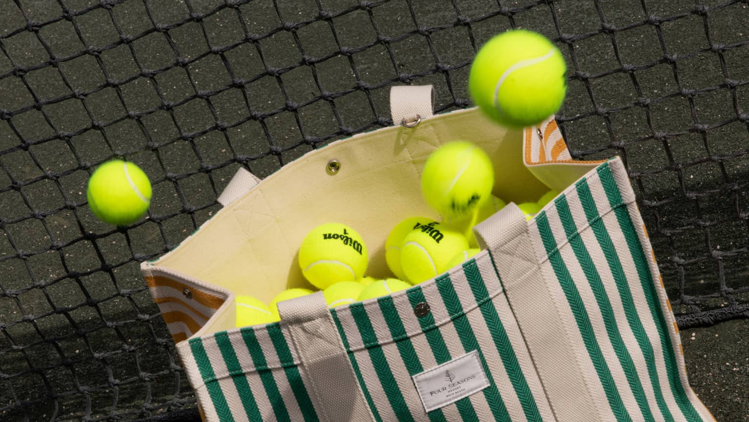 Green-and-white striped canvas bag filled with tennis balls leans against a net