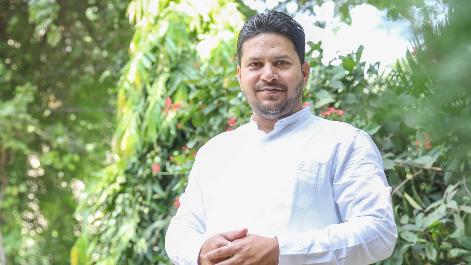 Person in long-sleeved white button-down stands with hands together outdoors in front of tree with red blooms