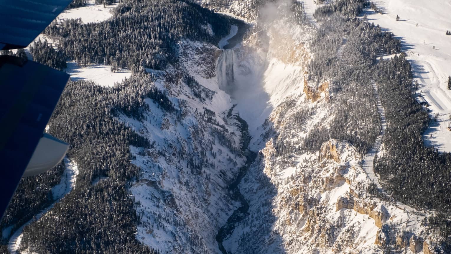 An aerial view of a waterfall plunging into a valley with sheer cliffs and a river, surrounded by trees and snow.