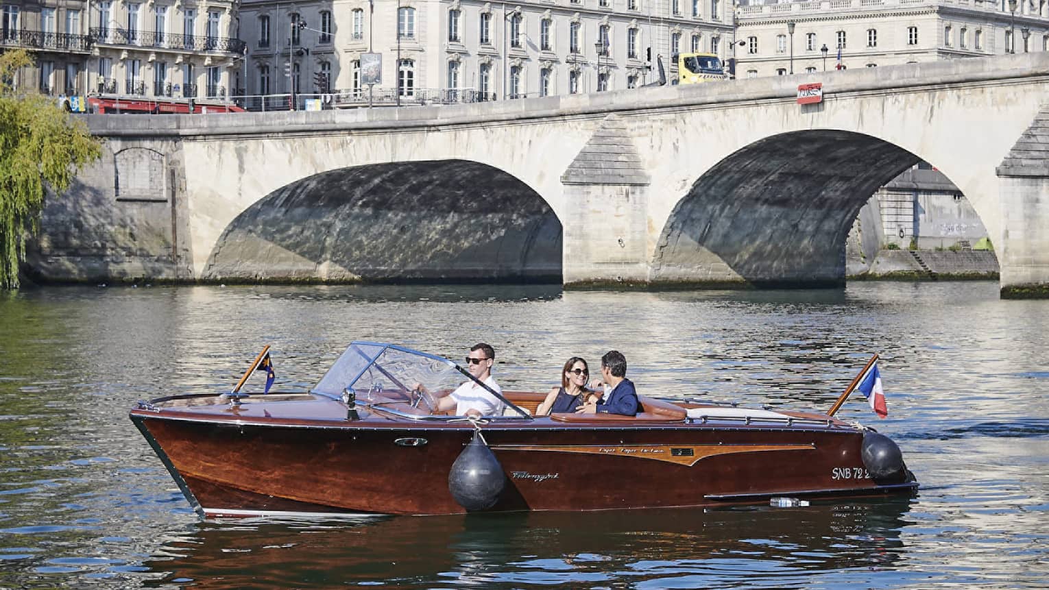 A couple relax in a brown speedboat cruising near a stone bridge along a river lined with white Haussmannian-style buildings.
