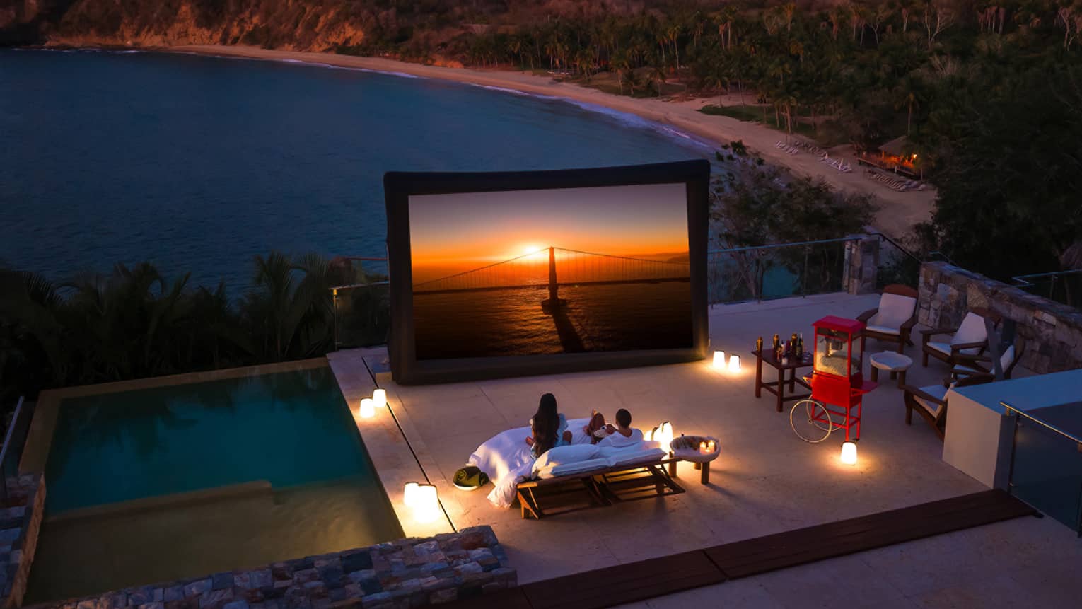 A couple watches a movie on a projector screen on a pool deck with a mountain view at sunset