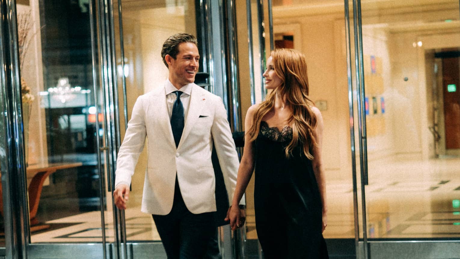A couple dressed in formal attire walks hand-in-hand in front of the glass doors of a modern building
