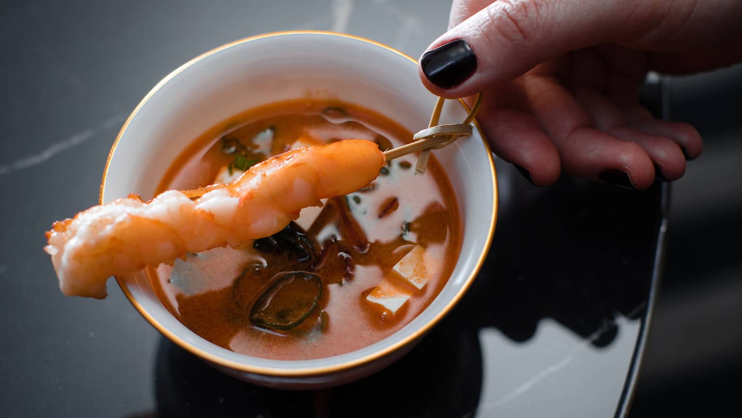 Hand holding cup of reddish-orange Miso Soup topped with prawn skewer over black marble table