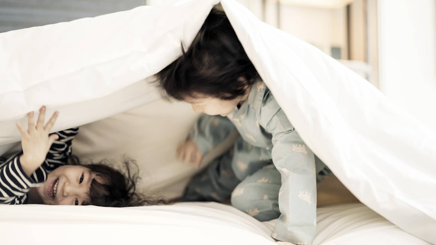 Two young children in pyjamas laugh, play under white blanket on bed
